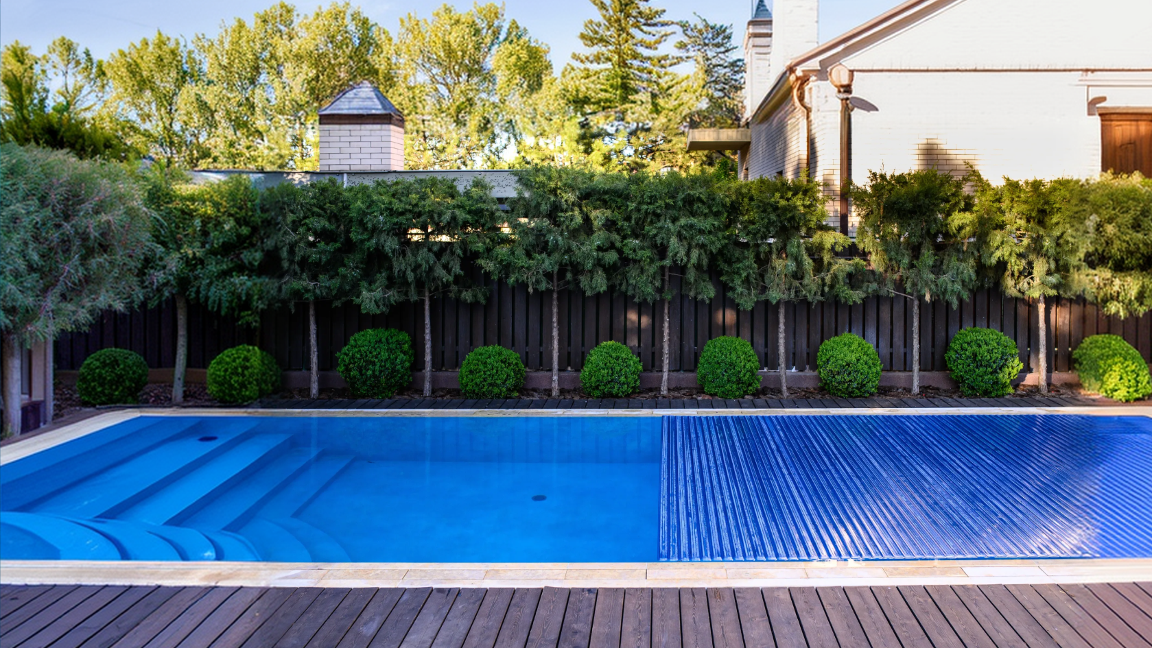 A covered outdoor swimming pool with a blue cover extending over a wooden deck, bordered by trimmed green shrubs and a dark fence, with trees and part of a house visible in the background.